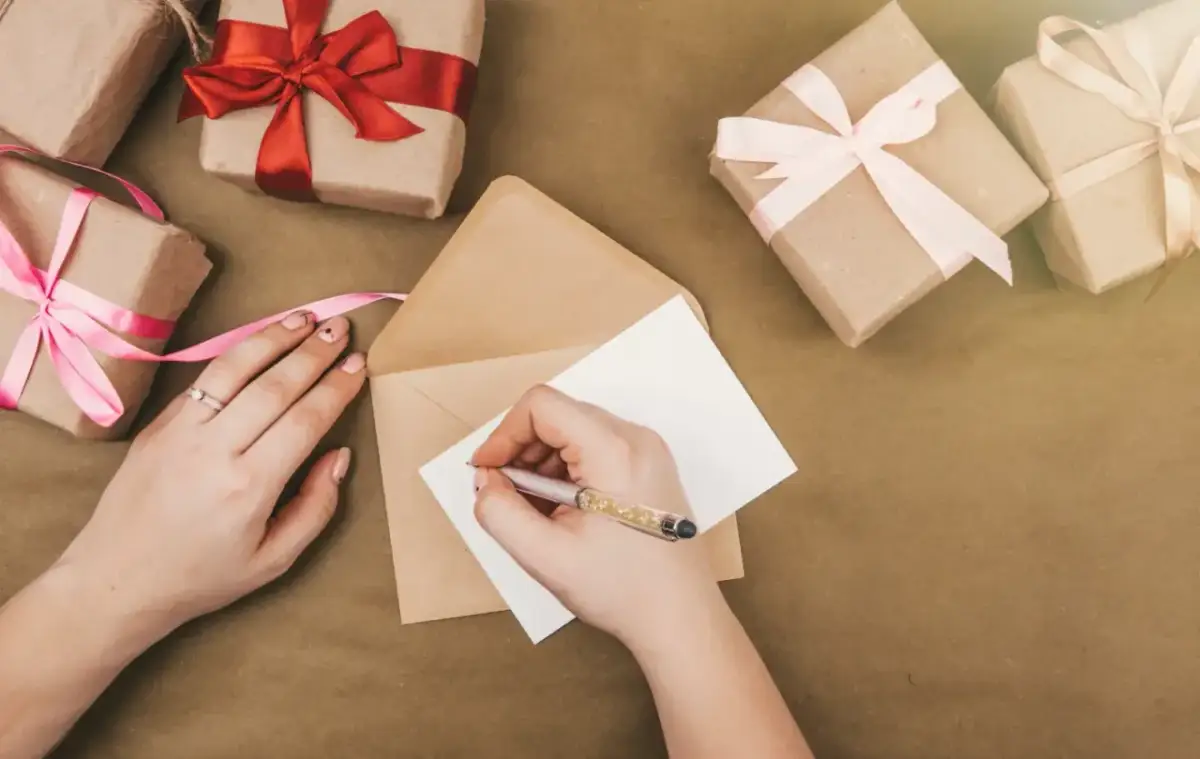 Dłonie piszą na kartce w kopercie, otoczone prezentami z wstążkami.