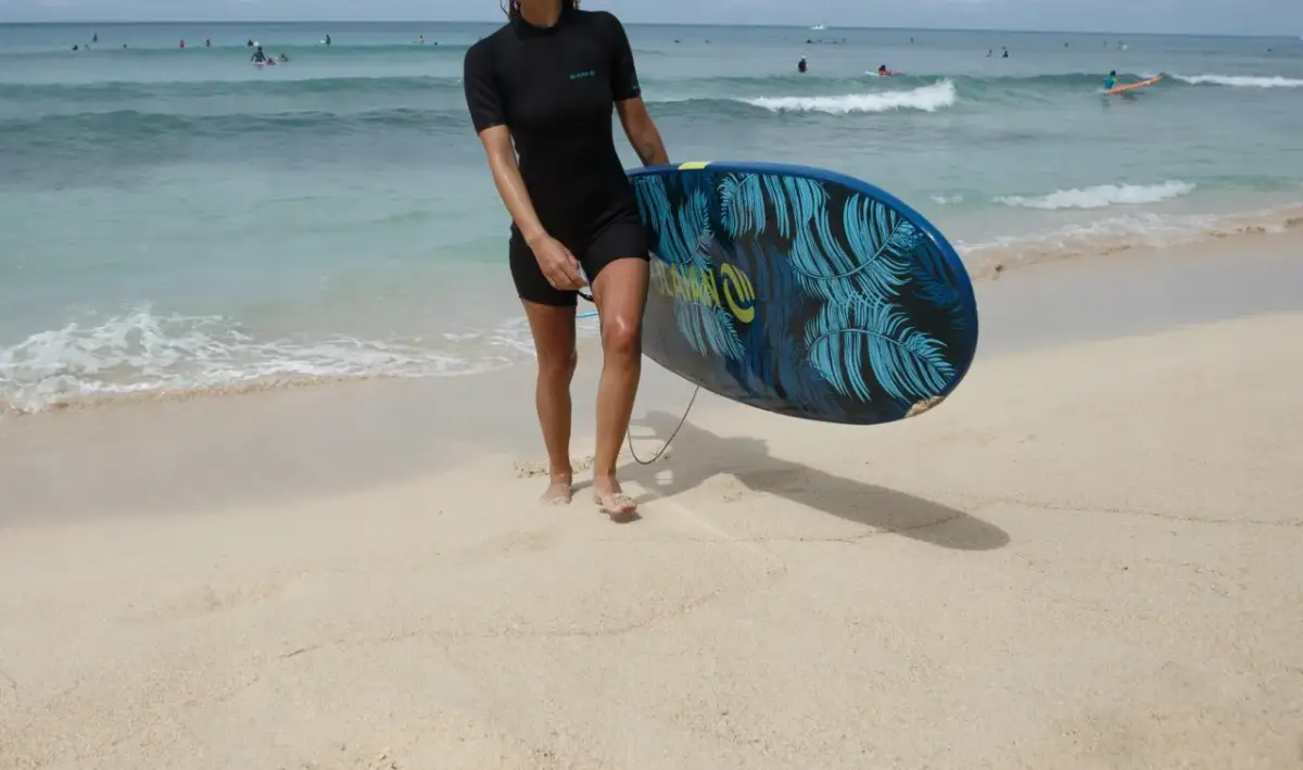 Kobieta w czarnej piance do pływania damskiej trzyma deskę surfingową z motywem liści palmowych na piaszczystej plaży.