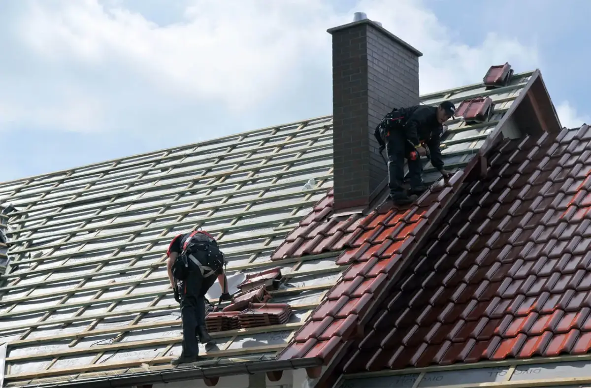 Dwaj dekarze układają dachówki. Widać rusztowanie i komin. Trudno ocenić ile dachówek na m2.