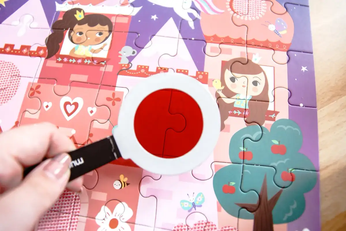 Kolorowa układanka dla dzieci z księżniczkami i zamkiem. Dłoń trzyma lupę, przez którą widać dwa czerwone elementy układanki.