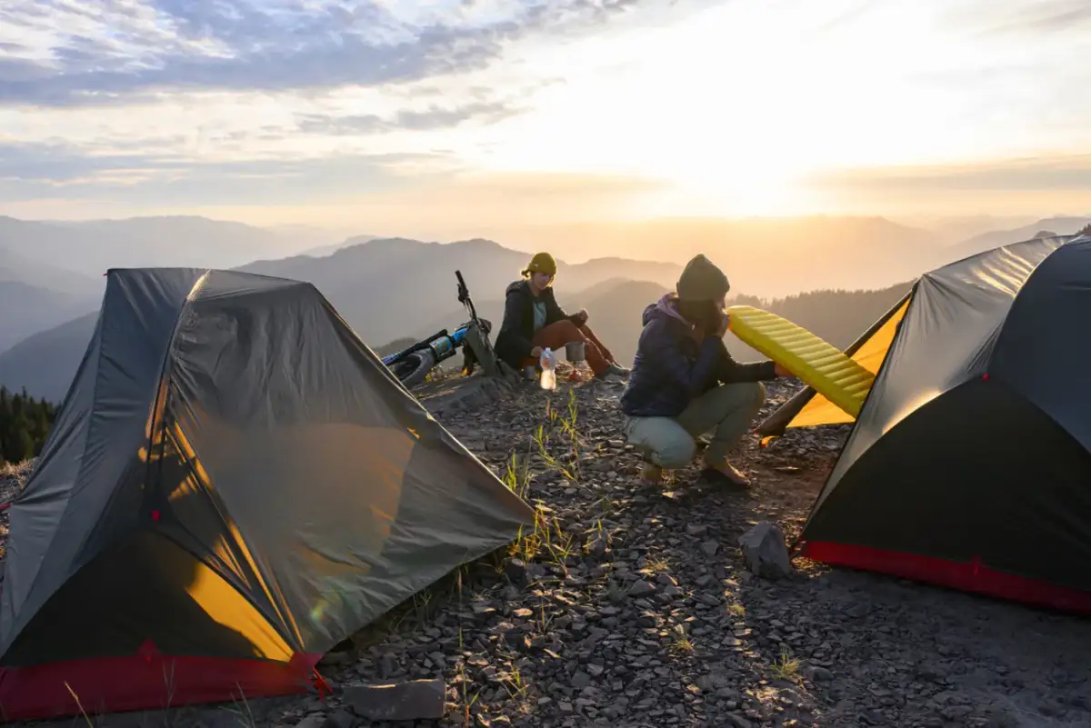 Dwie osoby przygotowują się do noclegu na szczycie góry. Jedna dmucha materac, druga siedzi obok roweru. W tle zachód słońca i góry. **Impregnacja namiotu** zapewni komfort.