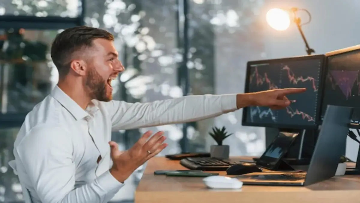 Młody mężczyzna z entuzjazmem analizuje wykresy giełdowe na monitorach. Chcesz wiedzieć, jak zacząć grać na giełdzie?