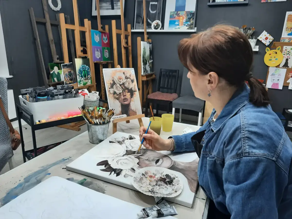 Kobieta w dżinsowej kurtce maluje obraz z kwiatami na płótnie w pracowni artystycznej.