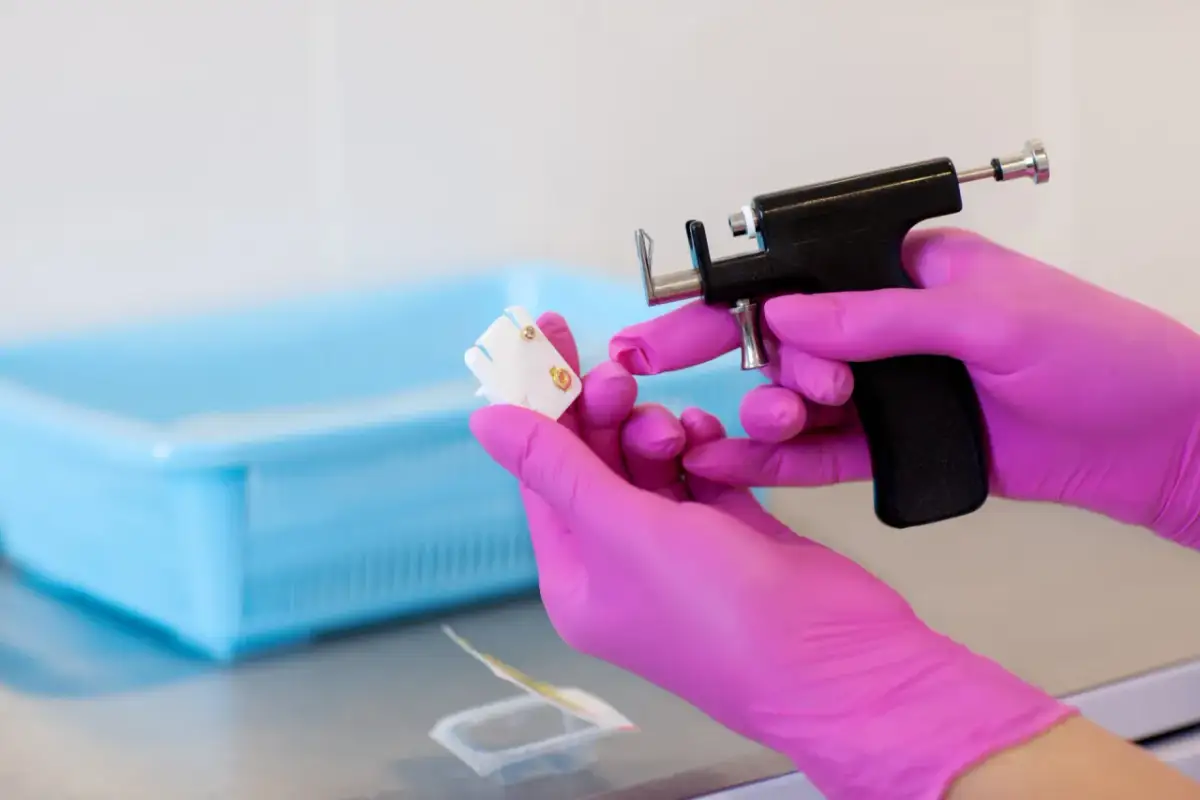 Ręce w różowych rękawiczkach trzymają pistolet do przekłuwania uszu i kolczyk.