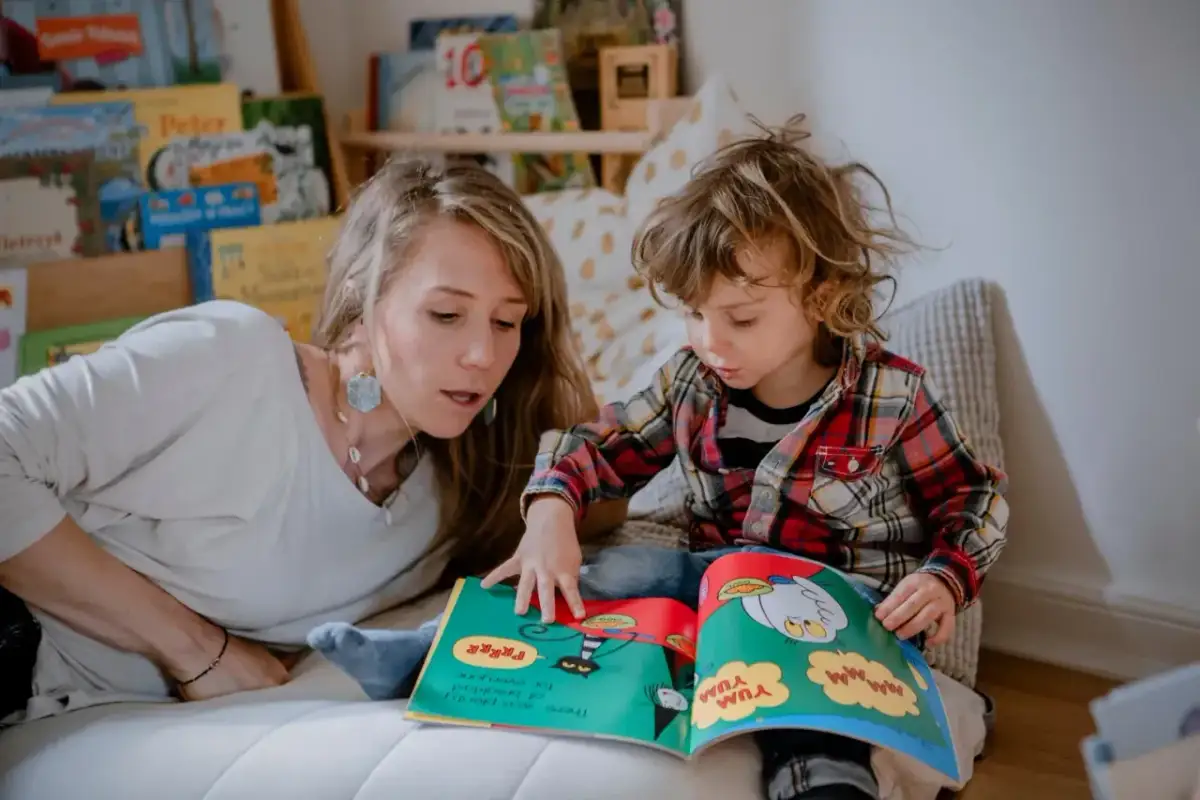 Kobieta czyta dziecku książkę, pokazując ilustracje. W tle regały z książkami, co oddaje atmosferę przytulnego miejsca do nauki i zabawy, jak wygląda żłobek.