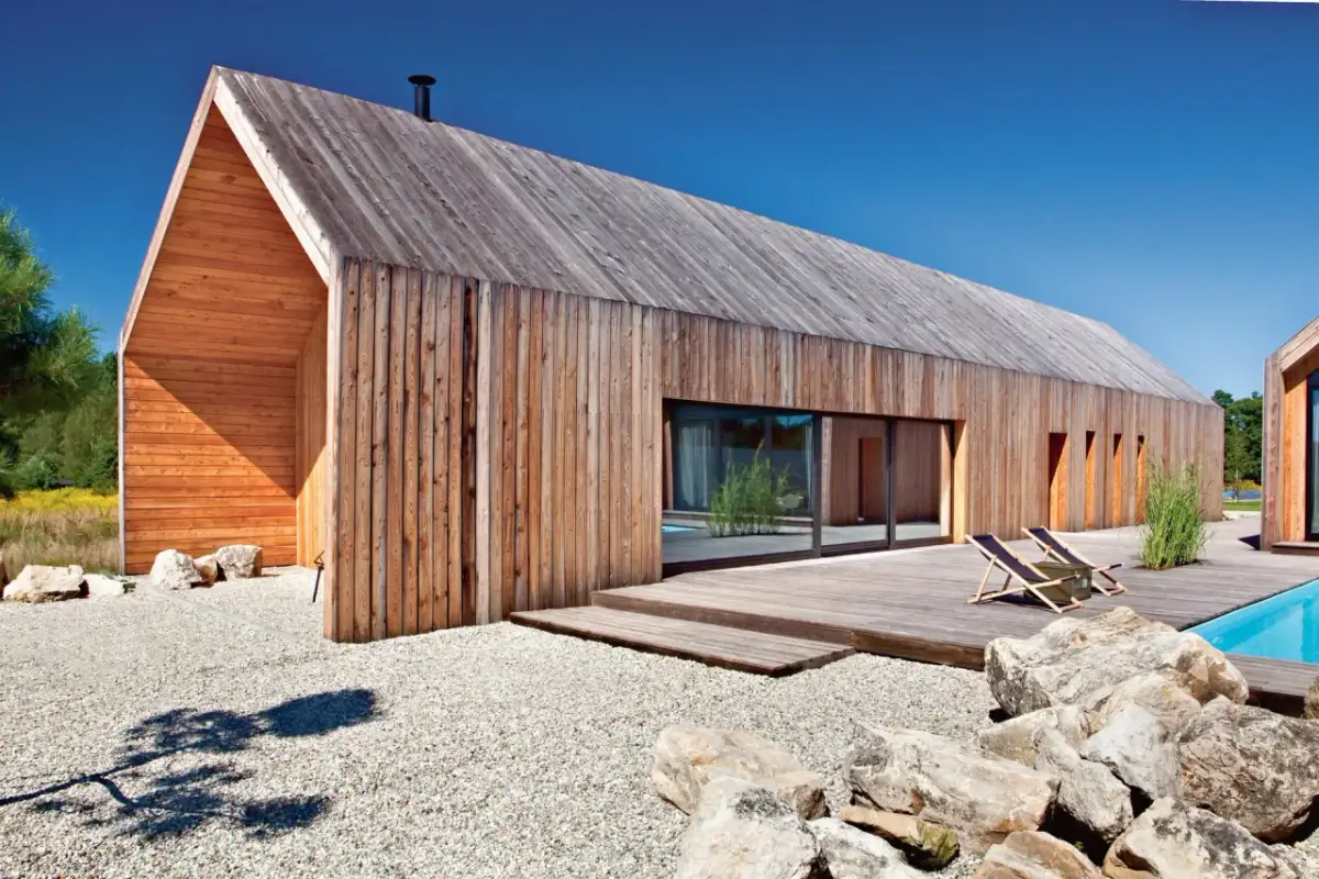 Nowoczesne technologie w budownictwie: drewniany dom z basenem i leżakami, otoczony kamieniami i żwirem.