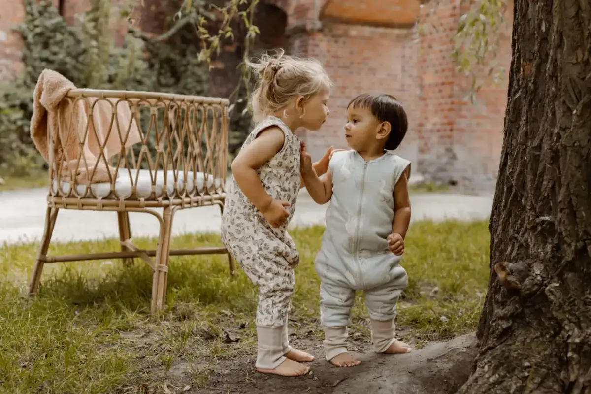 Dwoje dzieci w śpiworkach z nogami, jedno w jasny, wzorzysty kombinezon, drugie w błękitny z gęsiami, stoją przy drzewie.