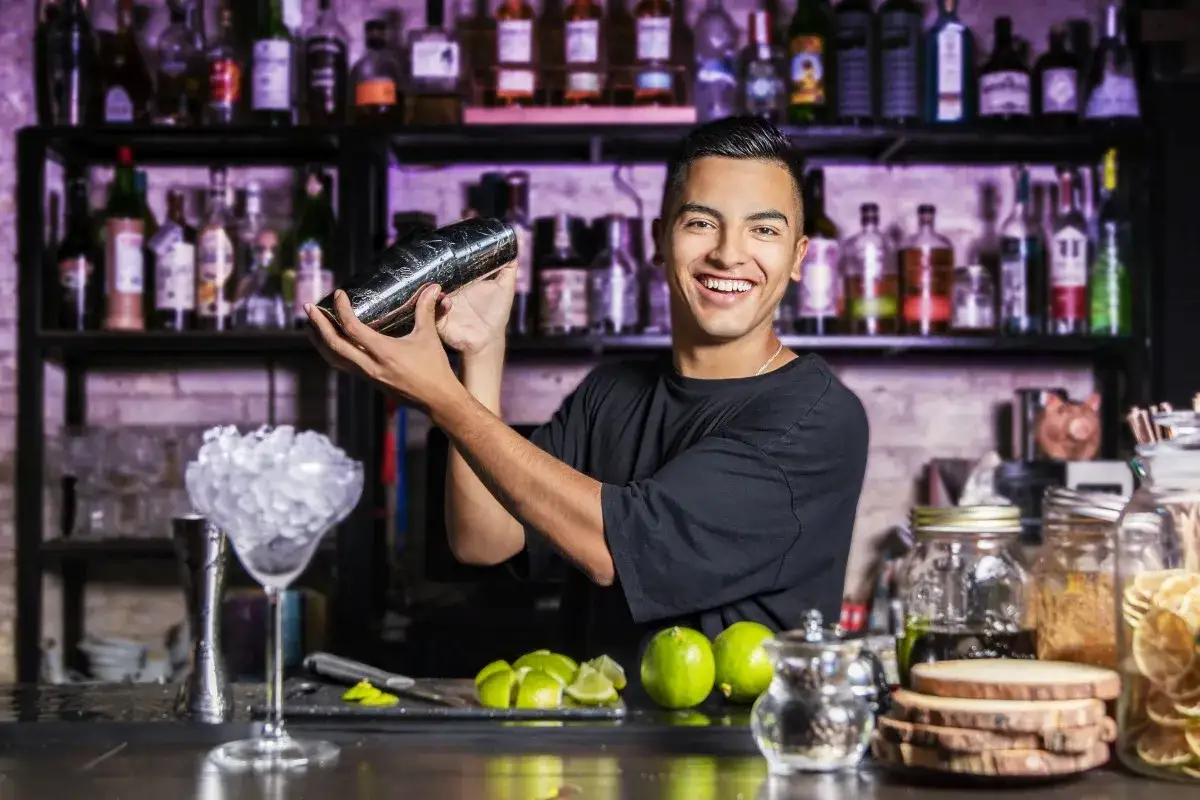 Uśmiechnięty barman miesza drinka w shakerze na tle półek z butelkami.
