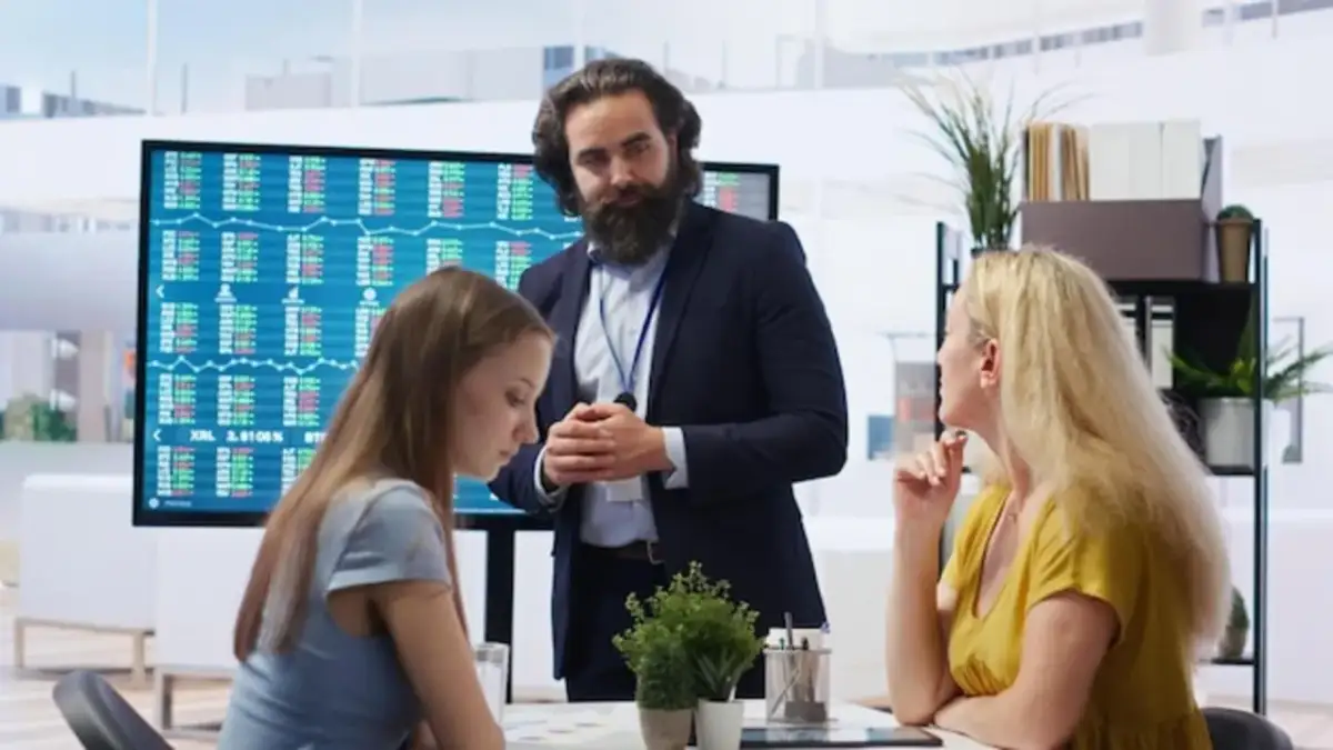 Szkolenie z giełdy dla bardzo początkujących. Mężczyzna w garniturze tłumaczy kobietom, jak zacząć inwestować, na tle wykresów giełdowych.