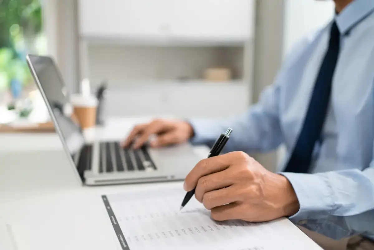 Mężczyzna w biurze pracuje przy laptopie i wypełnia dokumenty. Dowiaduje się, jak złożyć wniosek o wakacje kredytowe.