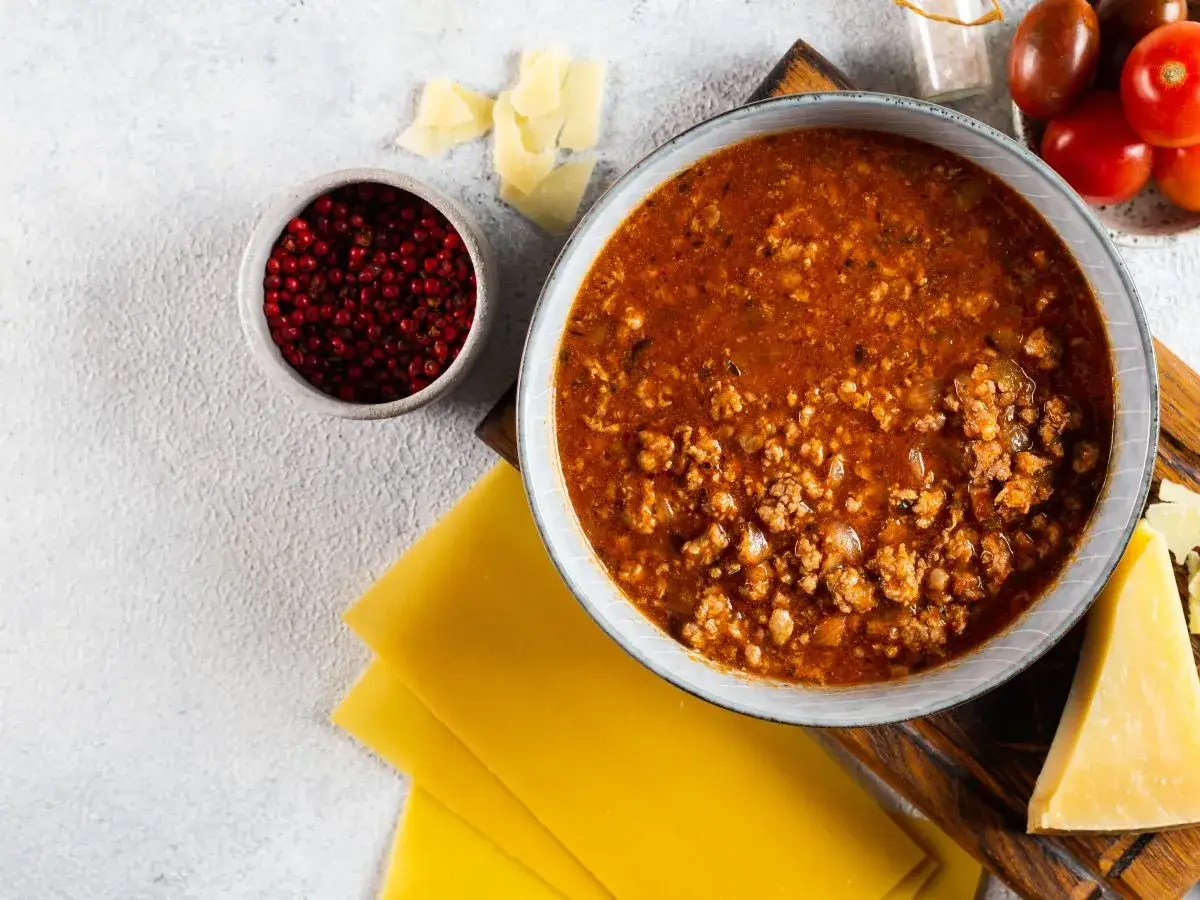 Sos bolognese w misce, obok płaty makaronu, parmezan, pomidorki i pieprz. Idealne składniki, by dowiedzieć się, jak zawekowac sos bolognese.