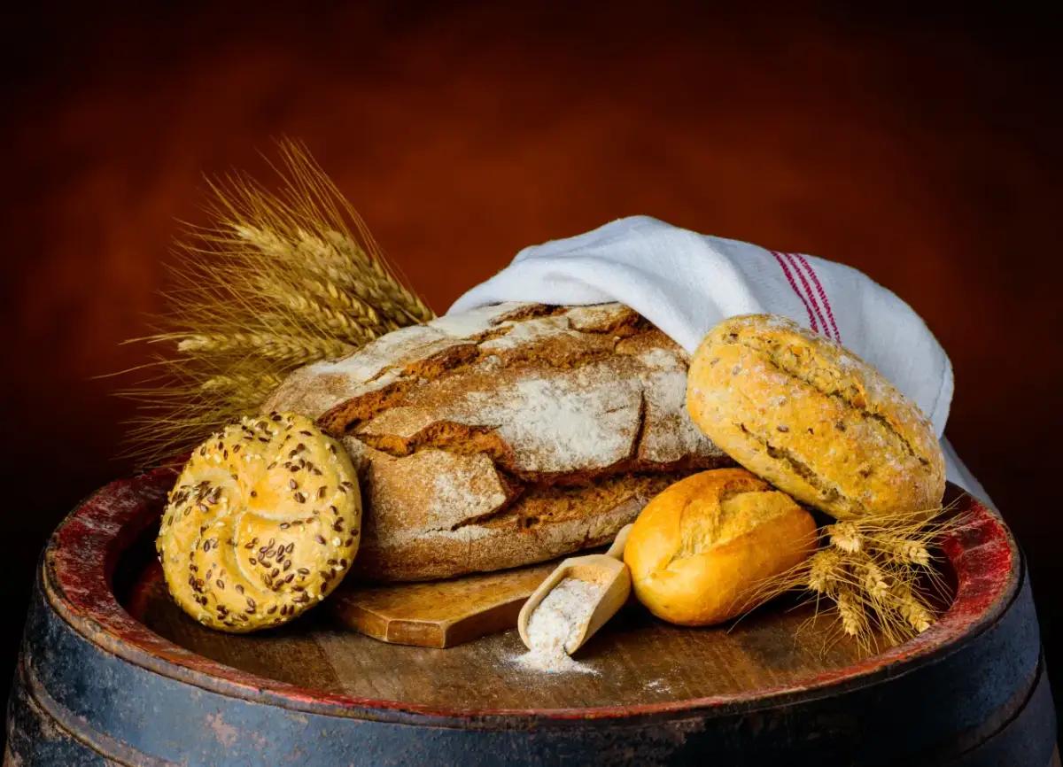 Na drewnianej beczce leży bochenek chleba, bułeczki i kłosy zboża. Czy chleb pszenny jest zdrowy? Zależy od jego składu.