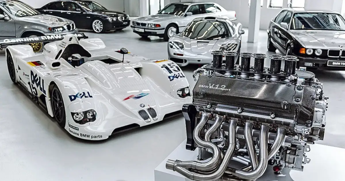 Silnik V12 BMW Motorsport obok białego bolidu wyścigowego i kilku klasycznych BMW.