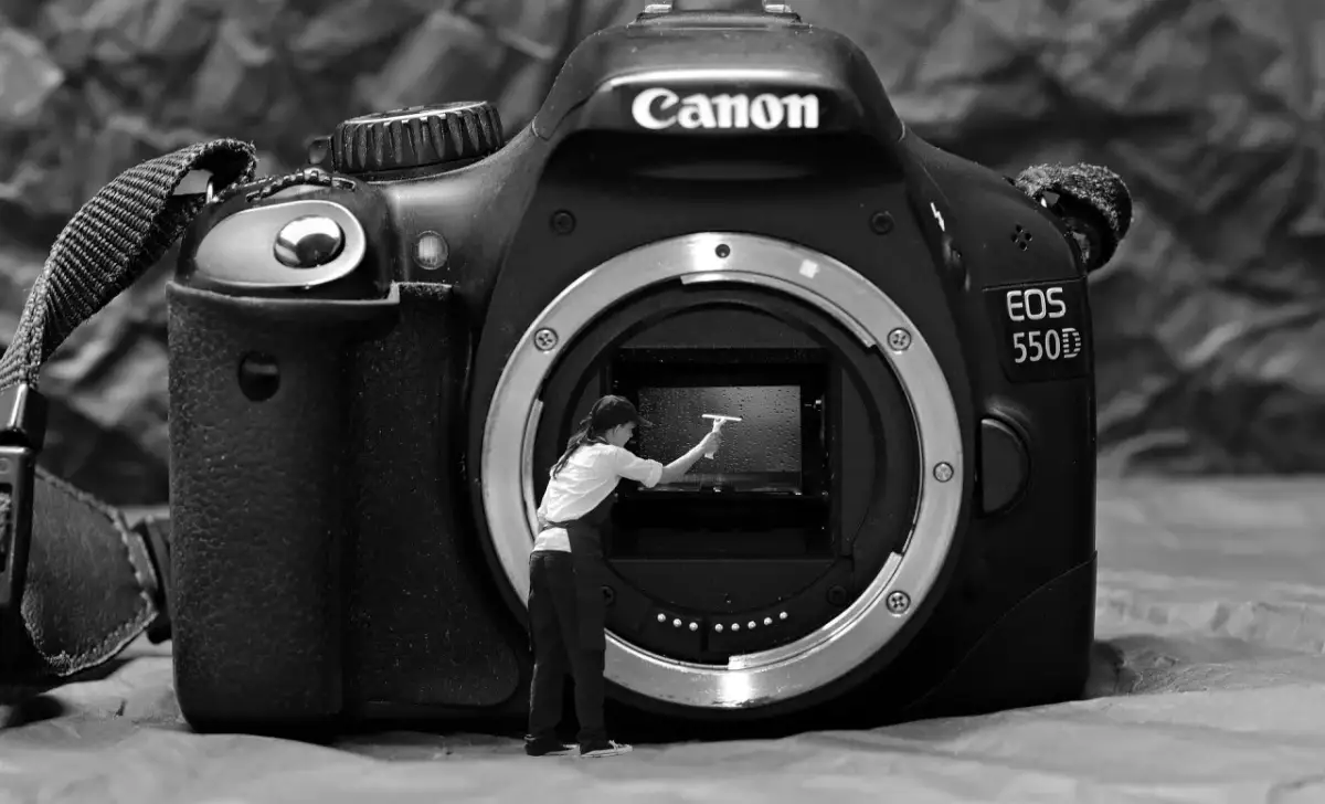 Miniaturowa postać czyści wnętrze aparatu Canon EOS 550D, pokazując jak poprawnie czyścić matrycę w bezlusterkowcu.