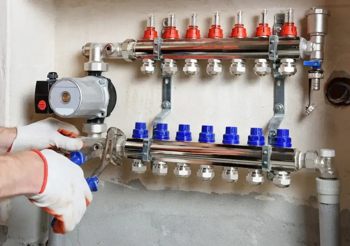 Instalator w rękawiczkach dokręca kluczem pompę obiegową do rozdzielacza ogrzewania podłogowego.