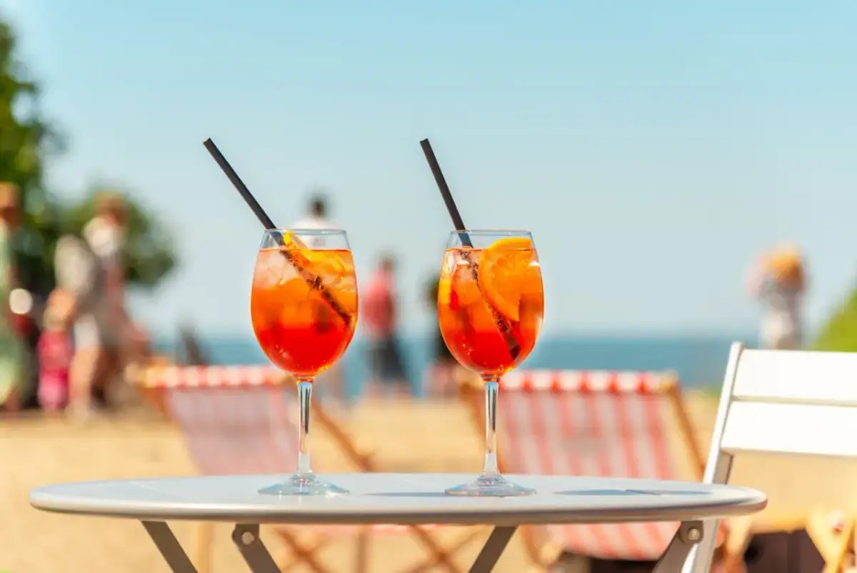 Dwa kieliszki Aperol Spritz z pomarańczą i lodem na stole, w tle plaża i morze.
