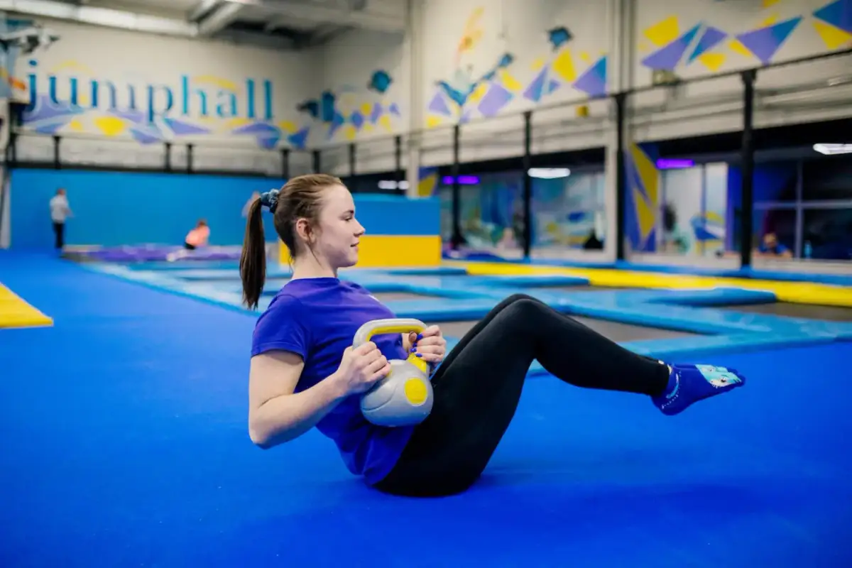 Kobieta ćwiczy z kettlebell na niebieskiej macie w sali gimnastycznej z trampolinami w tle.