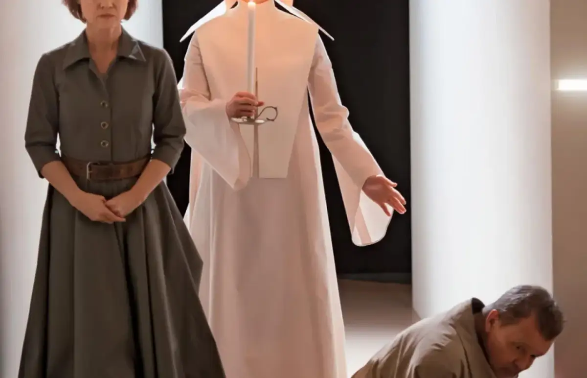 Scena z Teatru Narodowego "Jak być kochaną". Kobieta w szarej sukni, postać w białej szacie z świecą, mężczyzna klęczy.