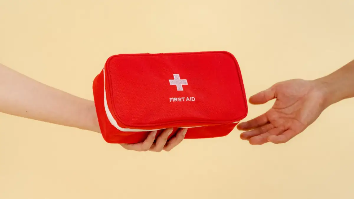 Czerwona apteczka z białym krzyżem i napisem "FIRST AID" jest przekazywana z ręki do ręki.
