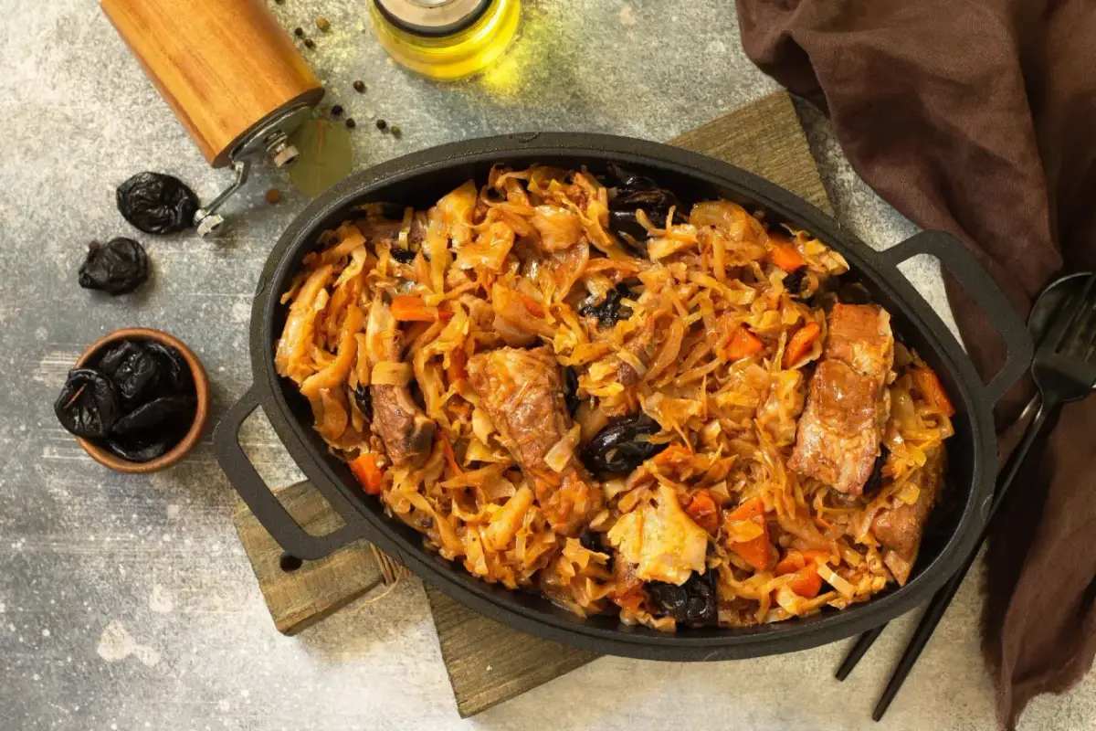 Pyszny bigos z kiszonej kapusty z mięsem i śliwkami, podany w żeliwnej patelni. Obok miseczka ze śliwkami i młynek do pieprzu.