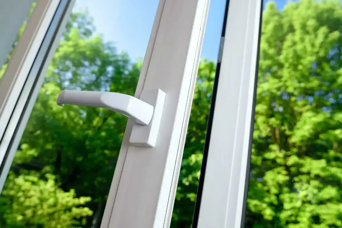 Uchylne okno z białą klamką, widok na zielone drzewa. Dowiedz się, jak zdjąć skrzydło z okna uchylnego.