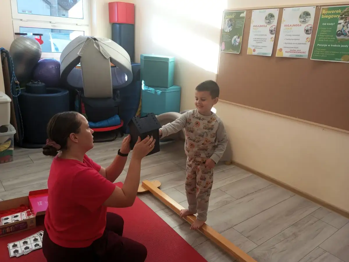 Fizjoterapeutka podaje chłopcu domek, który stoi na drewnianej belce.