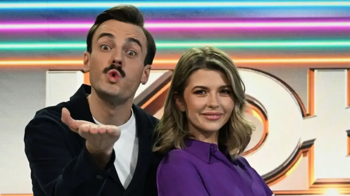 Agnieszka Dziekan, prowadząca Koło Fortuny, uśmiecha się do kamery. Obok niej stoi mężczyzna z wąsami.