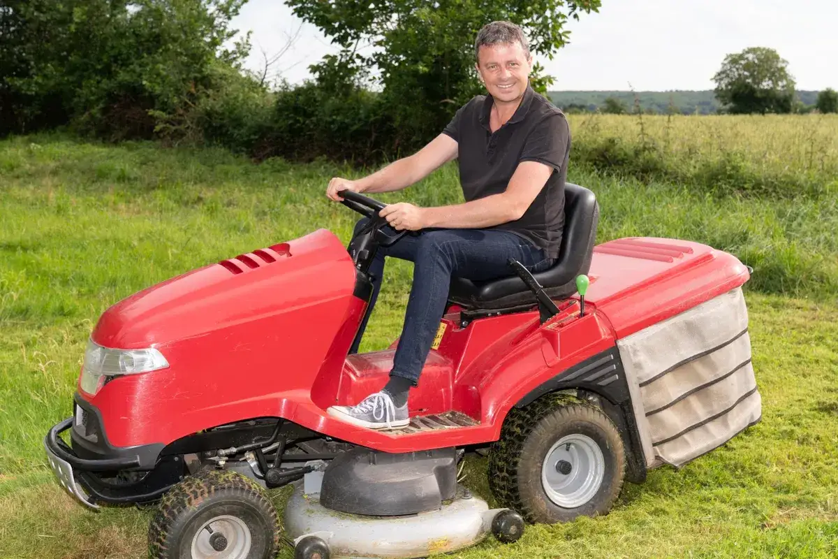 Uśmiechnięty mężczyzna siedzi za kierownicą czerwonego traktorka do koszenia trawy, gotowy do pracy w ogrodzie.