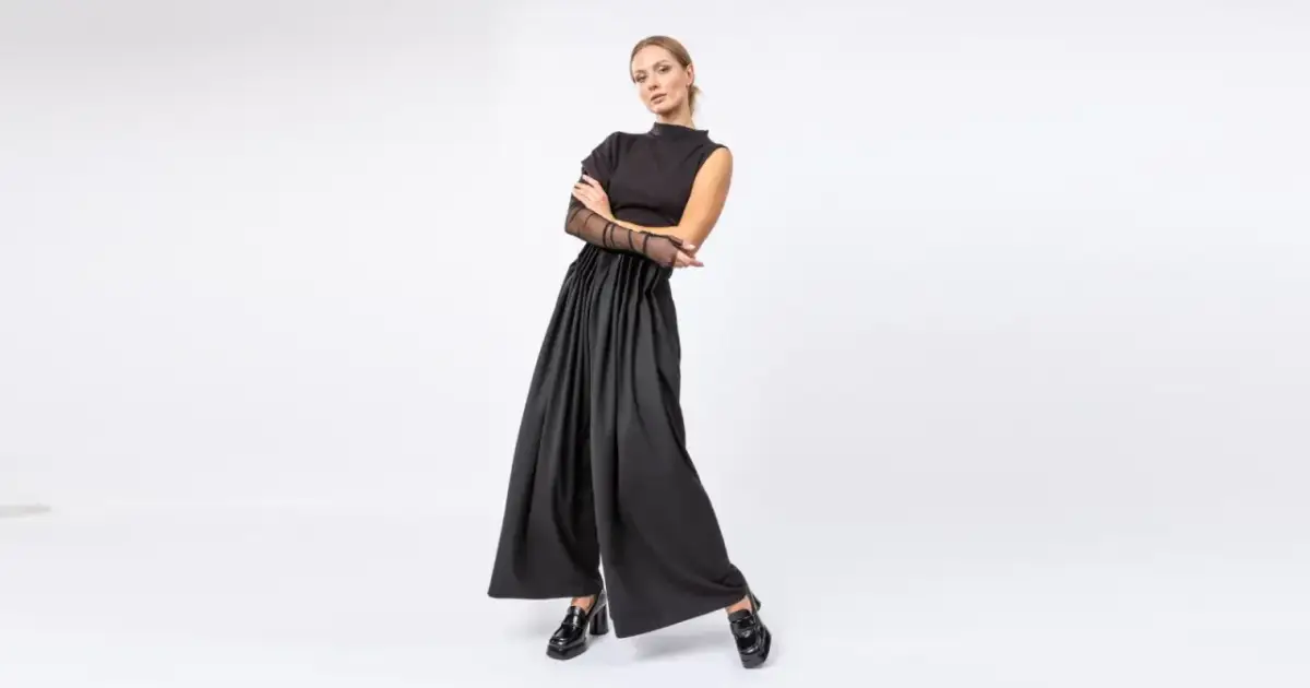 Kobieta w czarnym, eleganckim stroju z szerokimi spodniami i siateczkowymi rękawami, pozuje na białym tle.