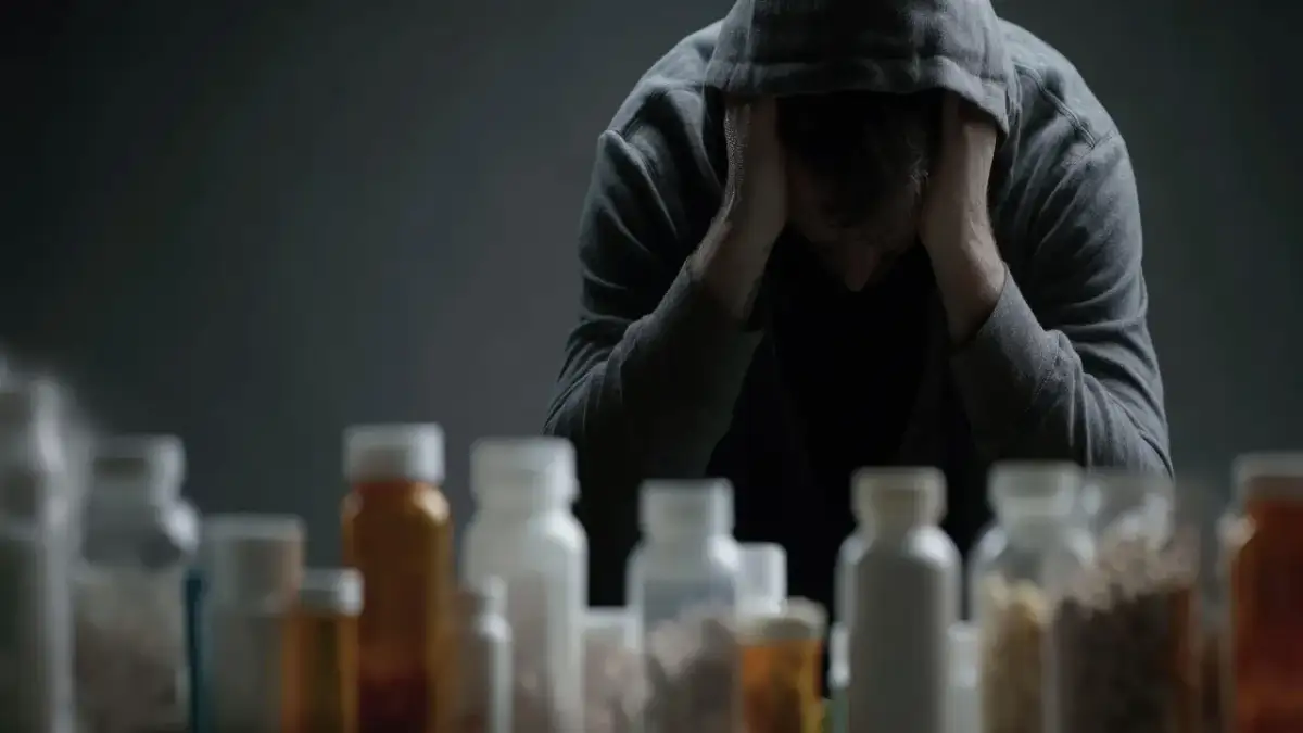 Zgarbiona postać w kapturze z rękami na głowie, przed nią rząd butelek z lekami.