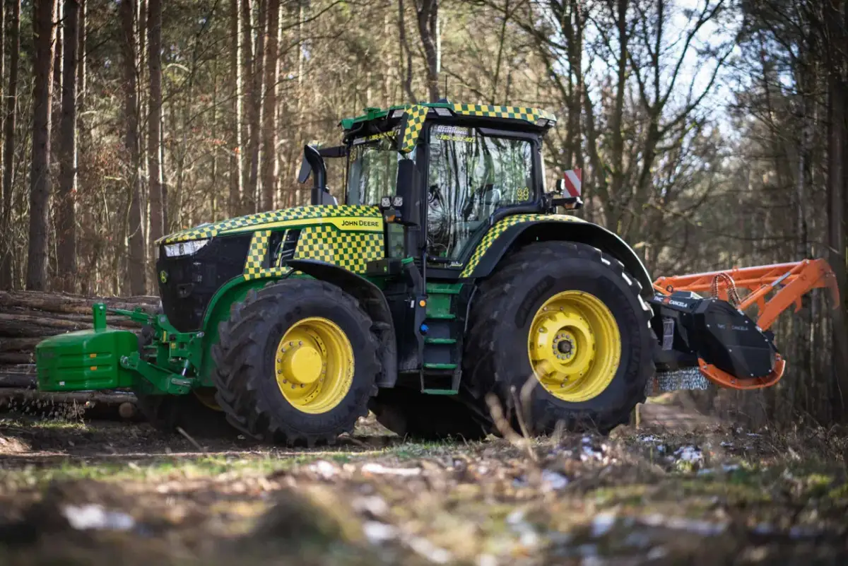 Traktor John Deere z charakterystyczną kratką w żółto-zielone wzory, wyposażony w mulczer do traktorka, pracuje w lesie.