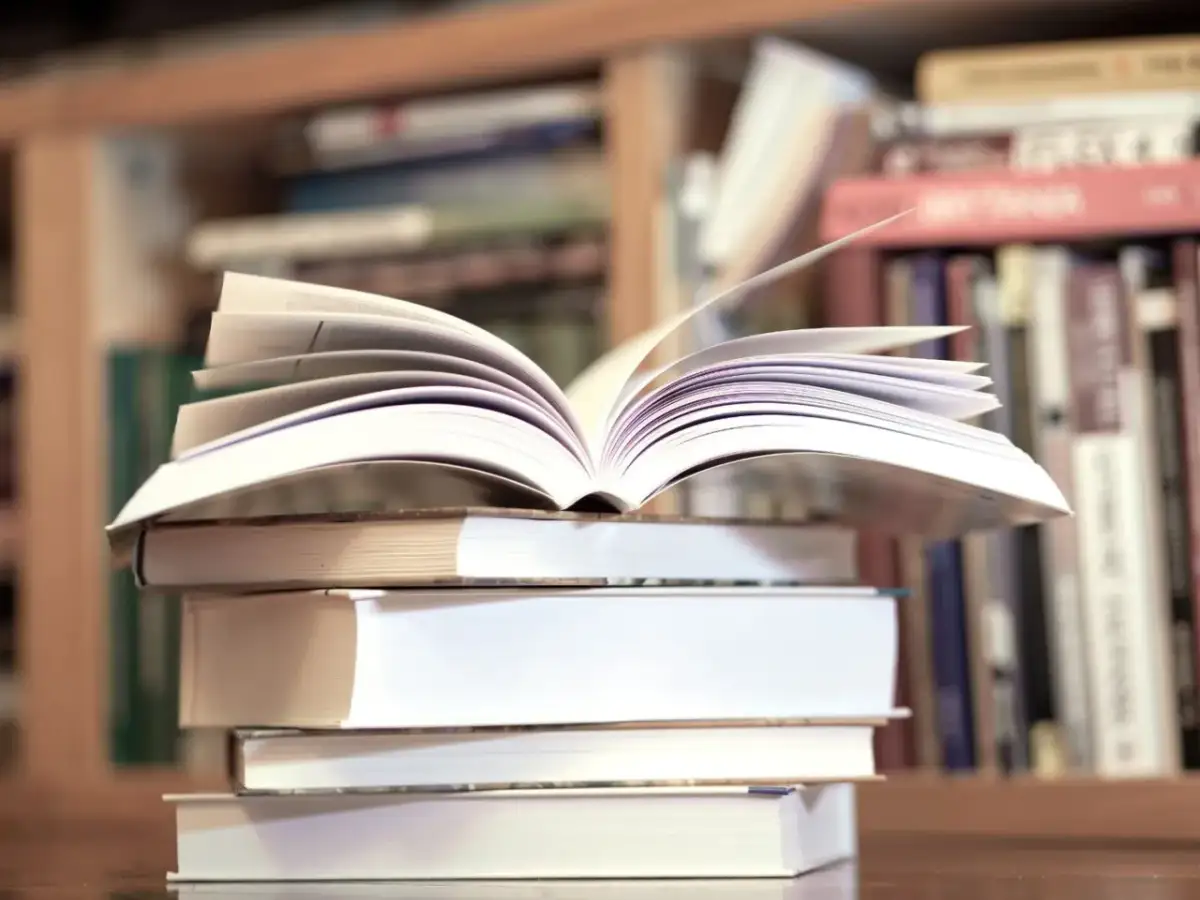 Otwarta książka na stosie innych, w tle regały pełne literatury, w tym klasyka jak "Zbrodnia i kara" gatunek.