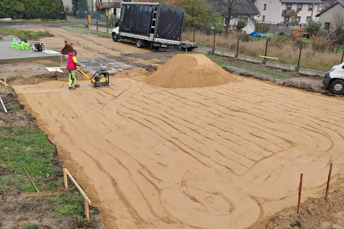 Pracownik zagęszcza piasek zagęszczarką. To kluczowy etap wymiany gruntu pod płytę fundamentową.