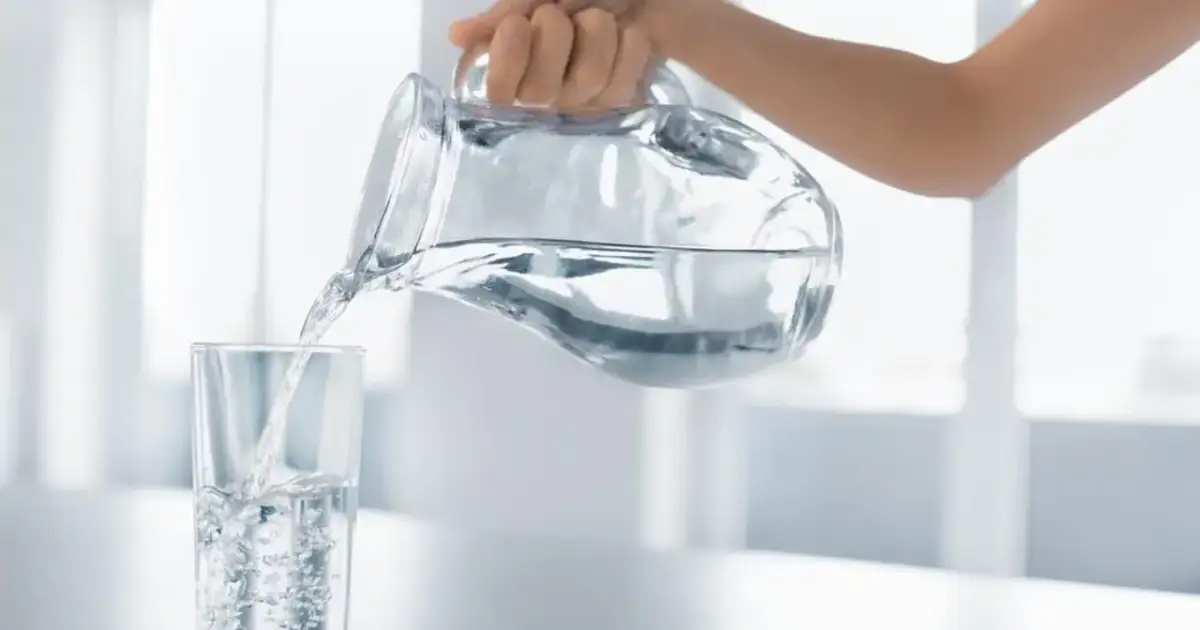 Dłoń przelewa czystą wodę alkaliczną z dzbanka do szklanki, tworząc bąbelki.