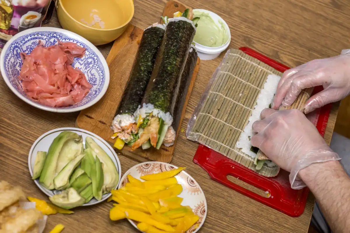 Ręce w rękawiczkach zwijają sushi na macie. Na stole składniki: ryż, nori, awokado, mango, imbir.
