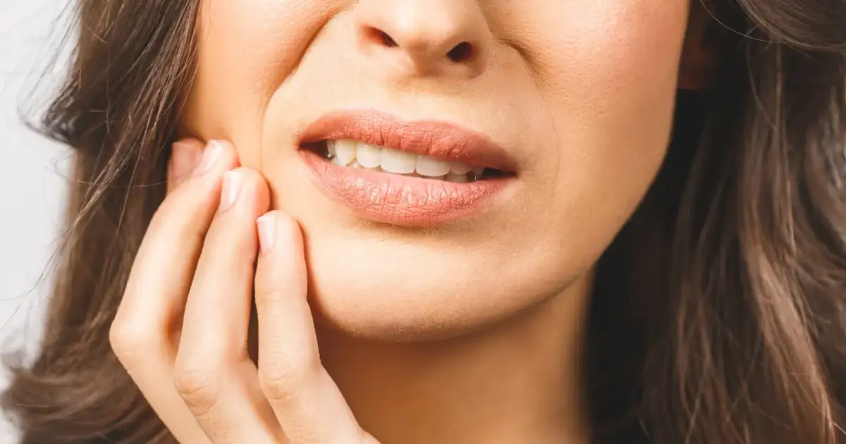 Kobieta trzymająca się za policzek, z widocznymi ustami i zębami.