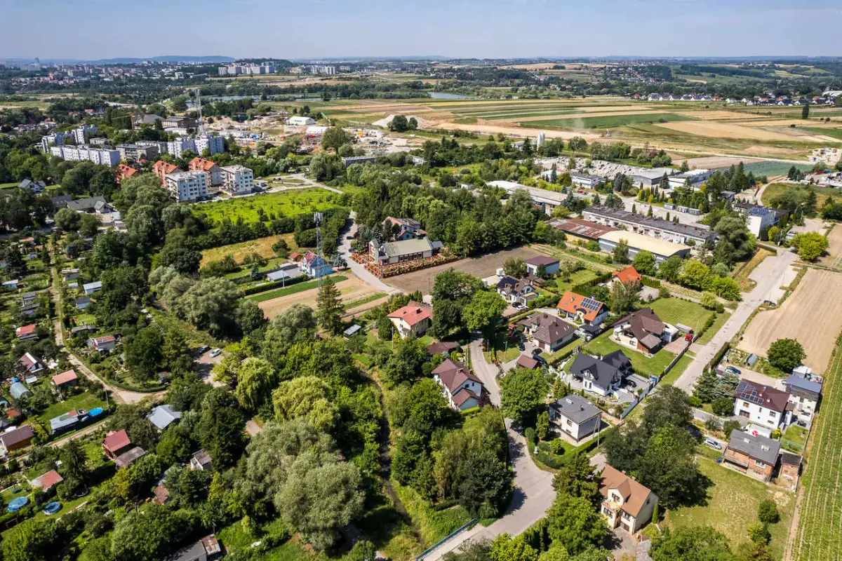 Osiedle domów jednorodzinnych na wzgórzach, otoczone zielenią i polami. W oddali widać zabudowania miejskie.