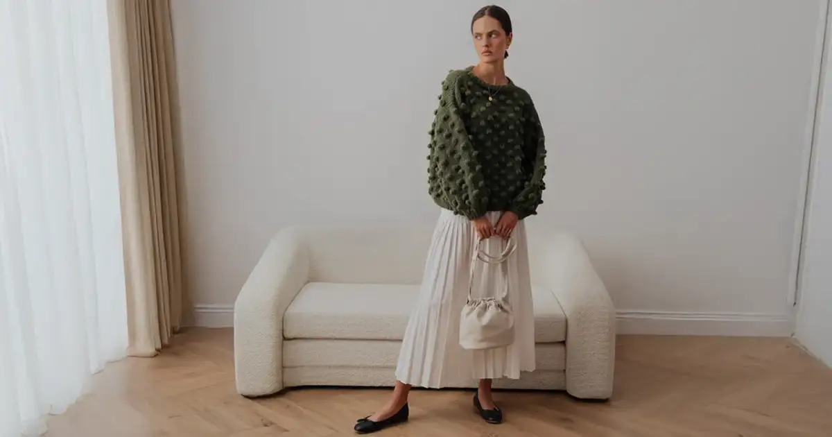 Zielony sweter z wypukłymi kulkami to idealna odpowiedź na pytanie, jaki sweter do letniej sukienki. Stylizacja z białą spódnicą i torebką.