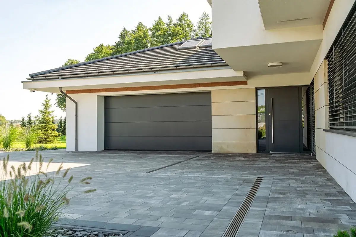 Nowoczesna brama garażowa, szara, segmentowa, z widocznymi panelami. Obok drzwi wejściowe.