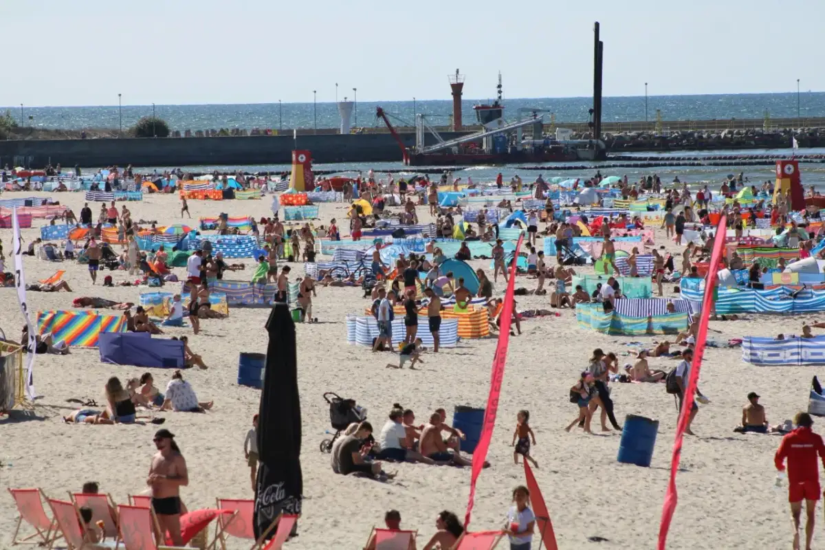Tłum ludzi na plaży w Łebie. W tle widać morze i port. Tyle mieszkańców ma Łeba, że plaża jest tak pełna.