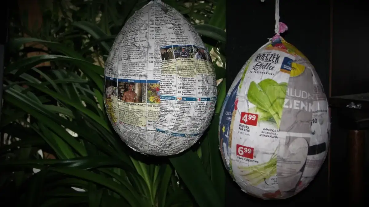 Jak zrobic jajko z gipsu w balonie? Dwa papierowe jajka, jedno z gazet, drugie z reklam, wiszą na tle zielonych liści.