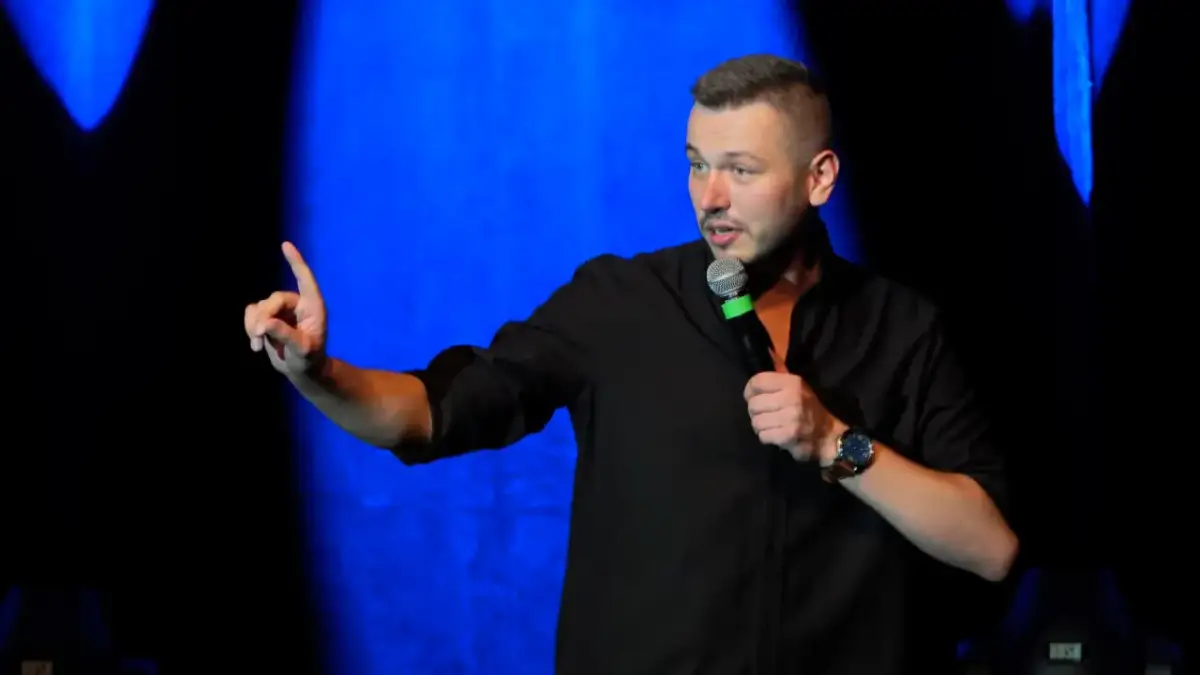 Mężczyzna w czarnej koszuli, trzymający mikrofon, gestykuluje na scenie. To jego top stand up.