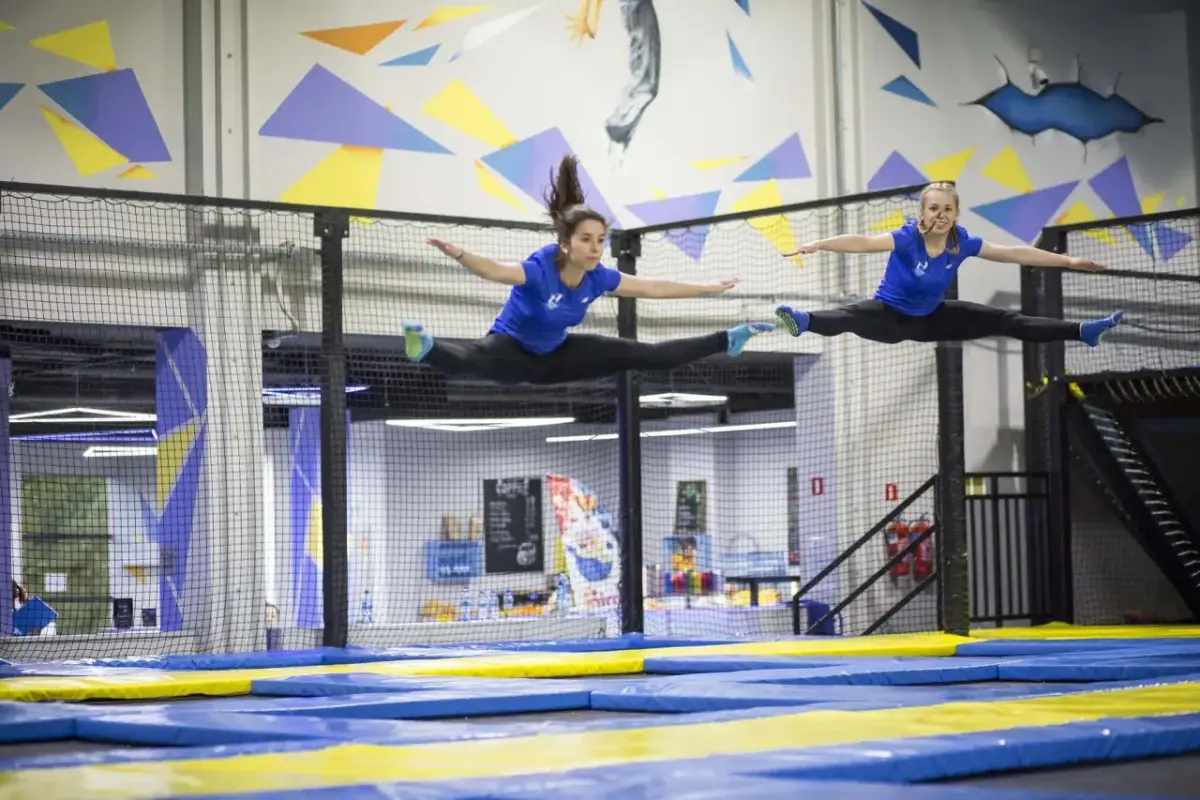 Dwie kobiety w niebieskich koszulkach wykonują szpagat w powietrzu na trampolinach.