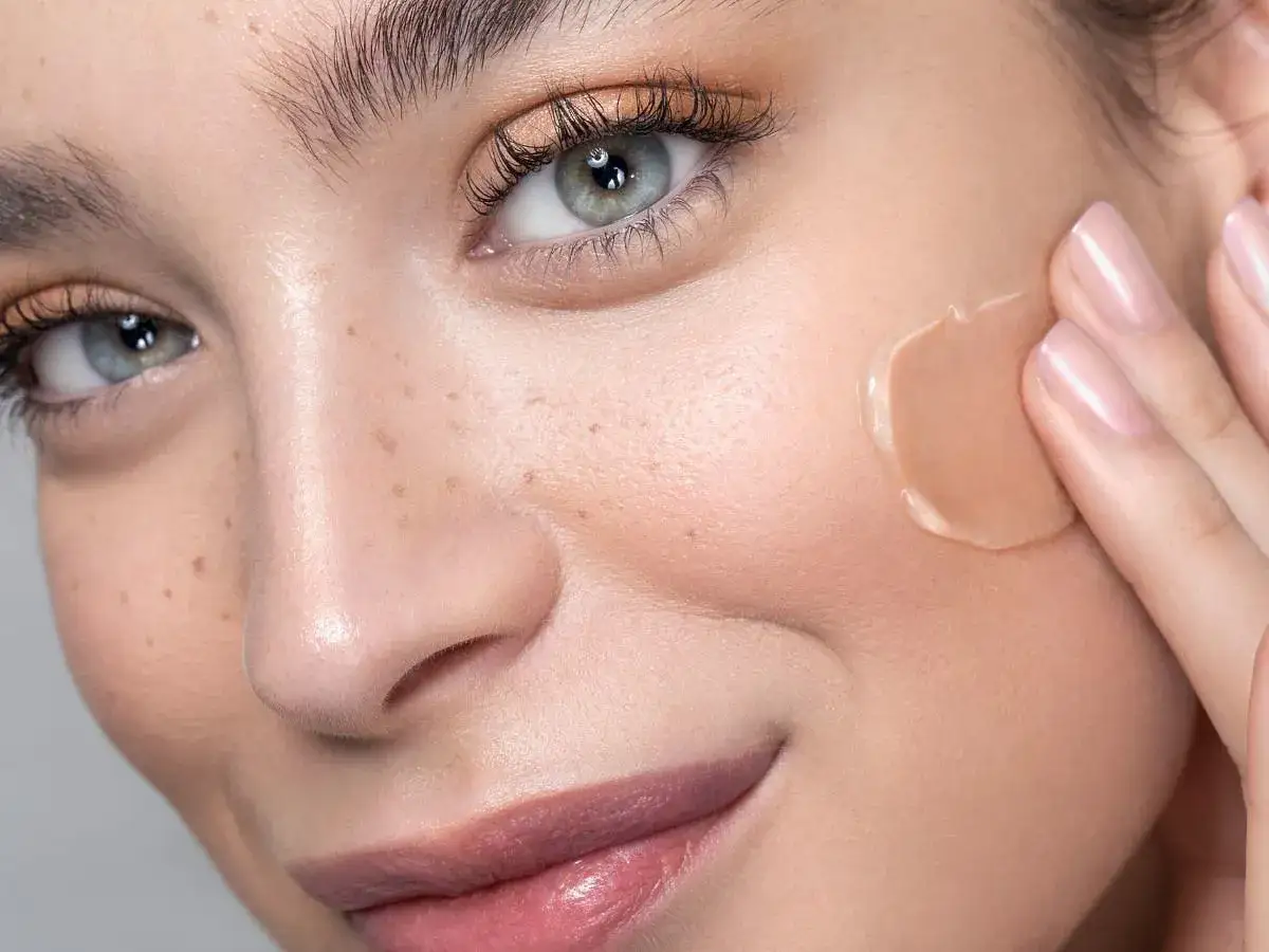 Zbliżenie na twarz kobiety z piegami, na policzku aplikowany jest krem BB.