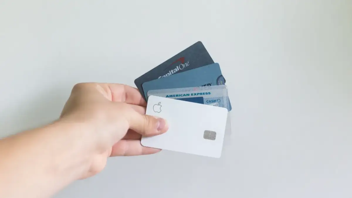 Dłoń trzyma kilka kart płatniczych, w tym Apple Card, American Express i Capital One. Standardowe wymiary karty bankomatowej są widoczne.