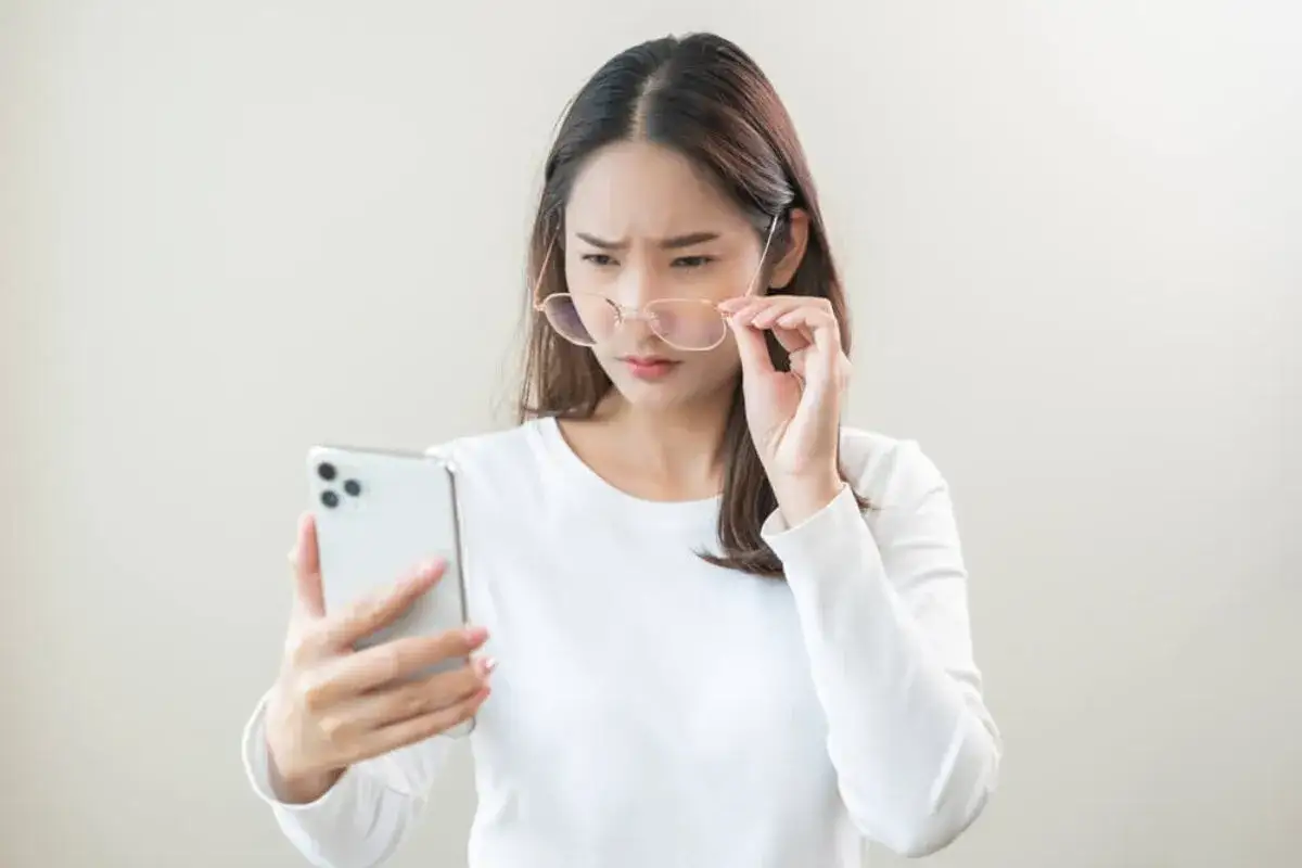 Kobieta w okularach z trudem patrzy na ekran telefonu.