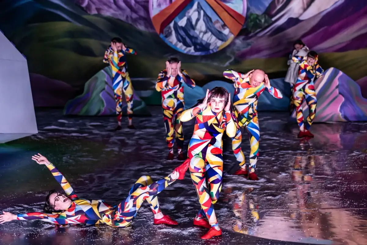 Artyści w kolorowych strojach tworzą syntezę sztuk w weselu, tańcząc na scenie z surrealistycznym tłem.