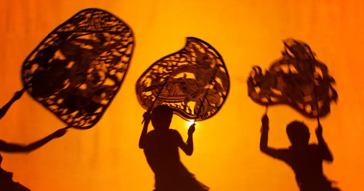 Trzy postacie z papierowymi lalkami tworzą cienie na pomarańczowym tle. Tak właśnie na czym polega teatr cieni – magia światła i wyobraźni.