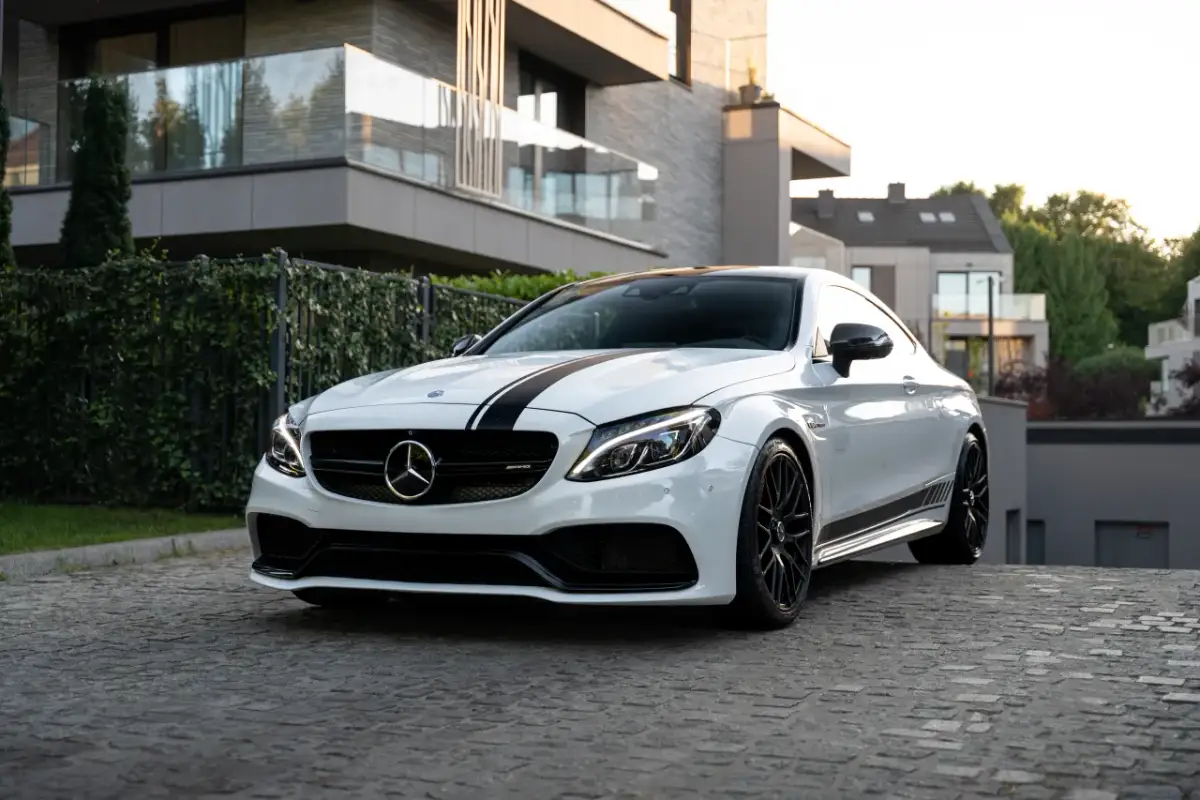 Biały Mercedes-AMG C63 Coupe z czarnymi pasami na masce, zaparkowany na brukowanej drodze przed nowoczesnym budynkiem.