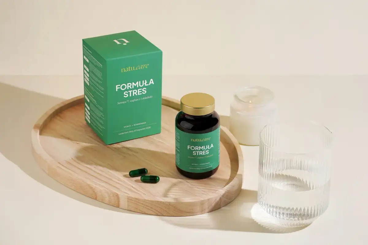 Zielone opakowanie i kapsułki suplementu diety "Formuła Stres" na drewnianej tacy, obok szklanki wody i świecy.